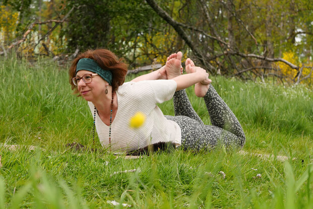 Ein freundliche Yogalehrerin mit roten Haaren macht Yoga in der Natur. Sie trainiert auf einer Wiese. Man sieht Bäume im Hintergrund. Die Yogaposition nennt sich Bogen.