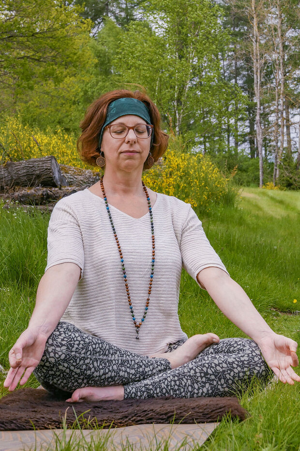 Ein freundliche Yogalehrerin mit roten Haaren macht Yoga in der Natur. Sie trainiert auf einer Wiese. Man sieht Bäume im Hintergrund. Sie sitzt in Meditationspose im Schneidersitz..